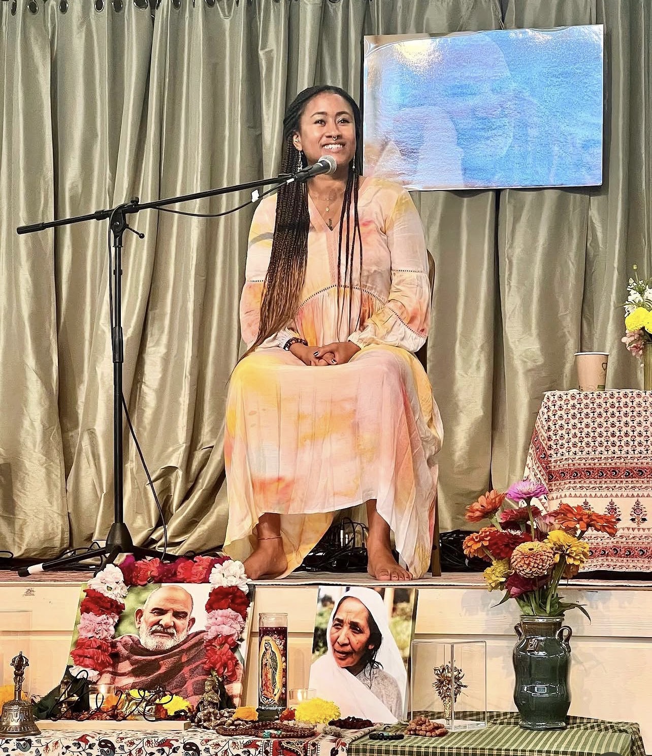 Dr. Sará King seated on stage offering a guided meditation, surrounded by flowers and an altar.