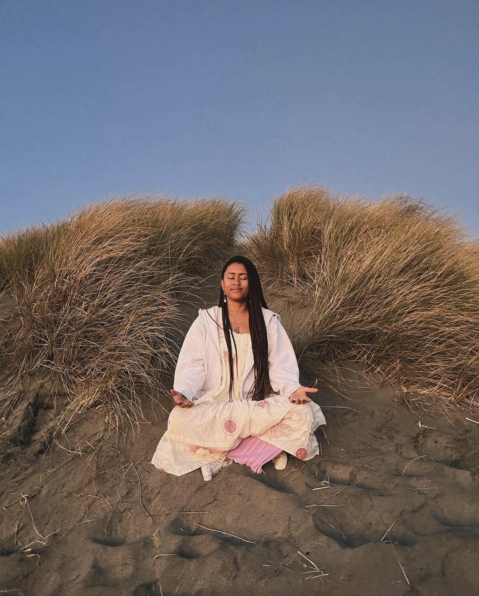 Dr. Sará King sitting in meditation on a coastal dune at Ocean Beach, San Francisco, surrounded by dune grass at golden hour.