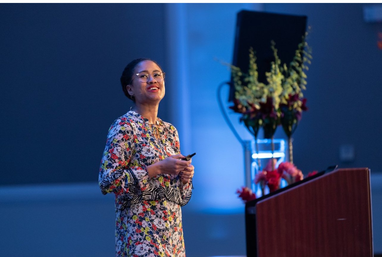 Dr. Sará King mid-sentence on a stage, microphone on, in a floral dress, soft blue stage lighting behind her, flowers at the podium.