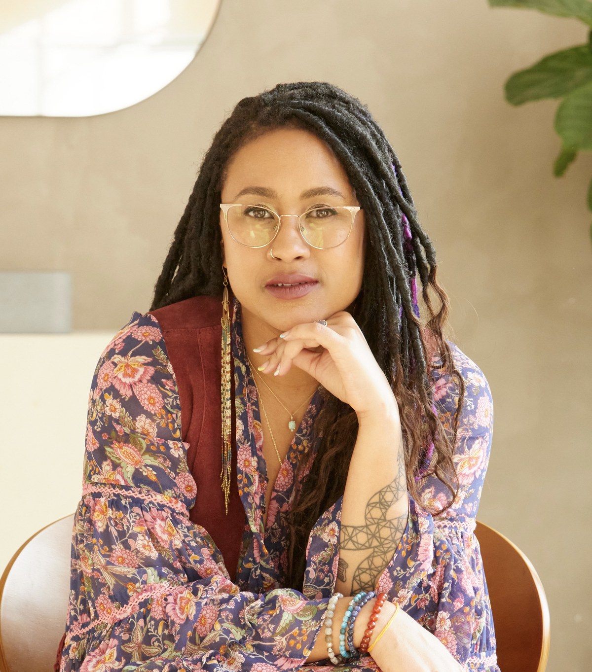 A studio portrait of Dr. Sará King in a floral kimono blouse over a burgundy vest, seated, chin resting on hand, eyes meeting the camera.