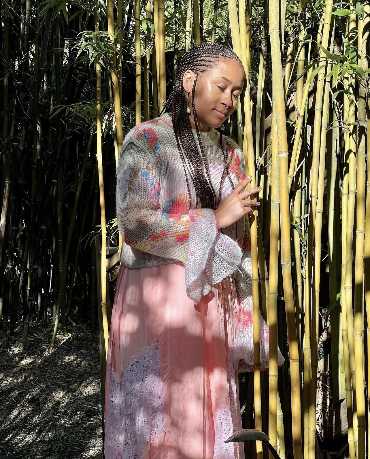 Sará King standing in a sunlit bamboo grove, wearing a soft floral knit sweater over a pink lace dress, eyes lowered, long braids resting over her shoulder.