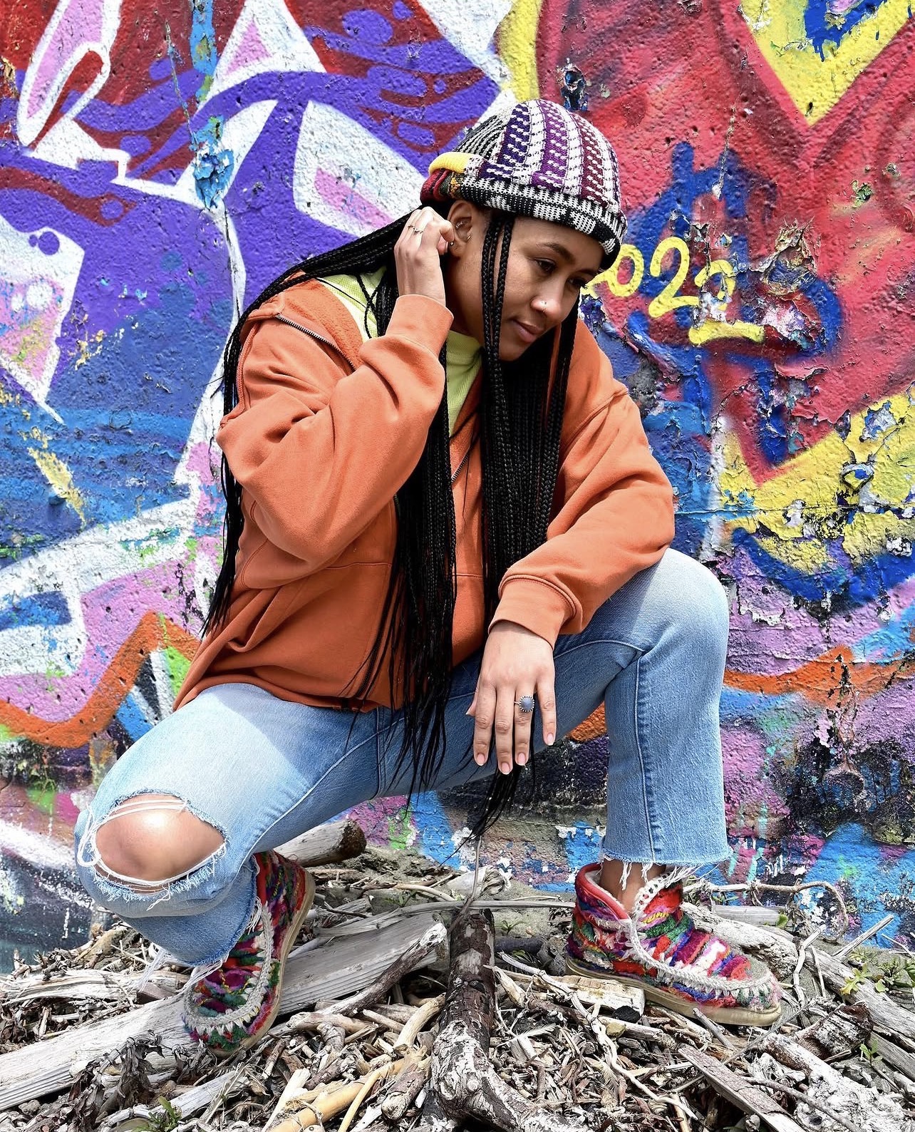 Sará King crouched on driftwood in front of a vibrant graffiti wall, wearing an orange hoodie and woven beanie, gaze low and contemplative.