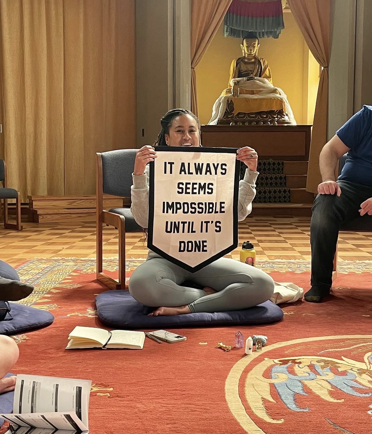 Sará King seated cross-legged on a meditation cushion in a Buddhist hall, smiling and holding a banner that reads &lsquo;It always seems impossible until it&rsquo;s done,&rsquo; a golden Buddha behind her.