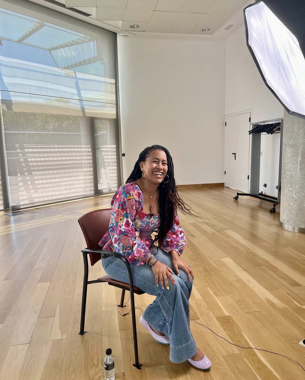 Sará King laughing on a chair in a sunlit studio with a softbox light, wearing a floral pink and blue top and blue jeans.