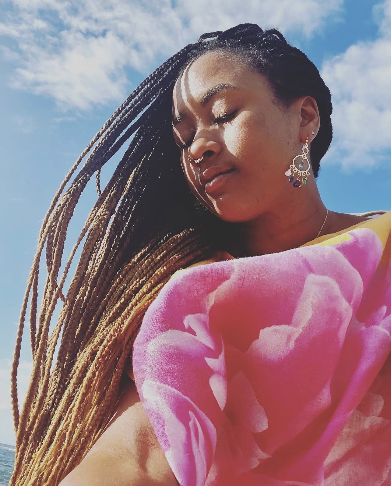 A close portrait of Sará King at the ocean — eyes lowered, long braids catching the sun, sky and sea behind her.