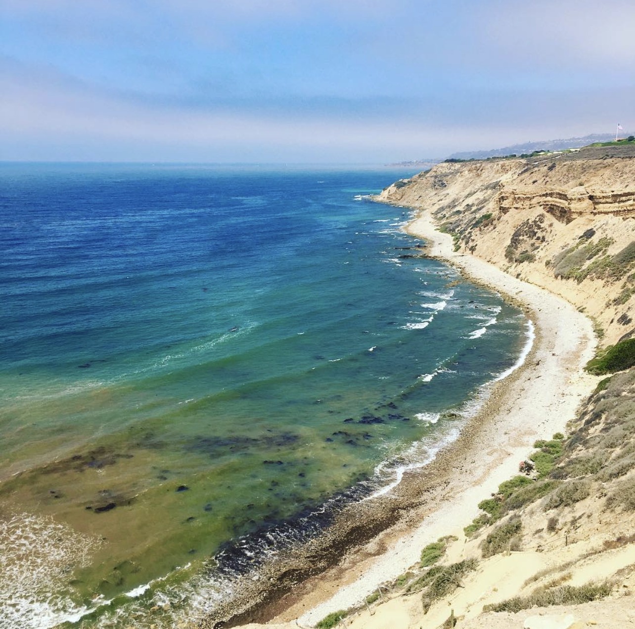 The Palos Verdes coastline — deep teal and cobalt Pacific water meeting sandstone cliffs under a hazy blue sky.