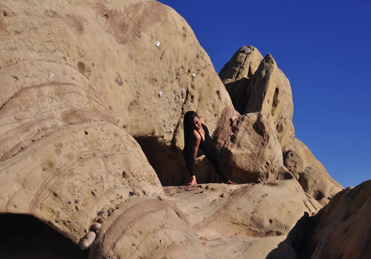 Sará King in a side-angle yoga pose against sun-warmed sandstone formations, long braids falling forward, deep blue sky behind her.